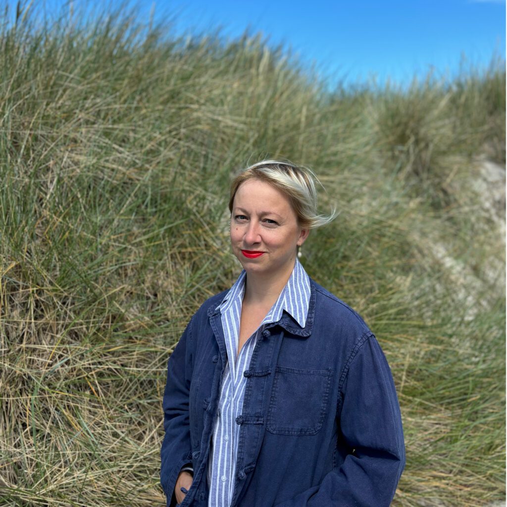 Vor einer grasbewachsenen Düne, über der der schmale blaue Himmel noch zu sehen ist, steht Frau Christl als Halbfigur. Ihre rechte Schulter ist leicht nach hinten gedreht, und ihr Blick richtet sich nach vorn. Sie hat halblanges blondes Haar, einen rot geschminkten Mund, trägt ein blau gestreiftes, oben geöffnetes Hemd und eine dunkelblaue Textiljacke. Ihre Hände sind in die Taschen der Jacke gesteckt.